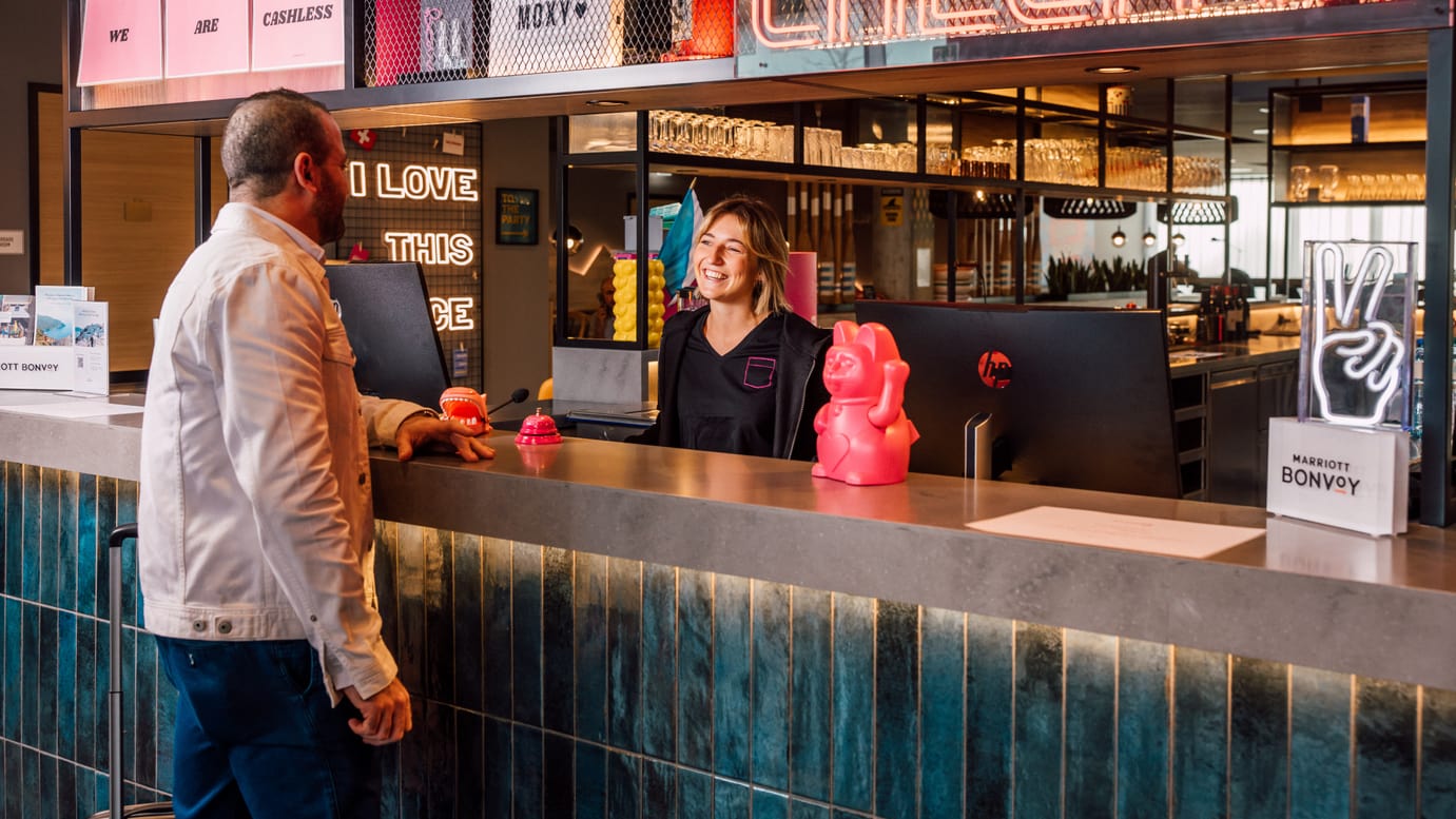 Mann am Hotelempfang mit lächelnder Rezeptionistin und neonfarbenem Dekor.