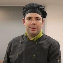 Porträt eines Mannes in Kochuniform mit schwarzer Kochmütze und grünem Schal.