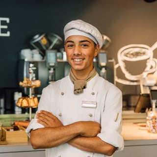 Junger Bäcker in Uniform, verschränkte Arme, lächelt in einer Bäckerei.