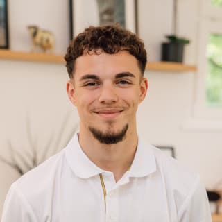 Junger Mann in weissem Polohemd, lächelnd in einem Büro.