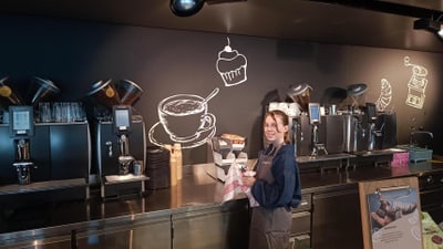 Frau steht hinter Cafétheke mit Kaffeetafeln im Hintergrund.