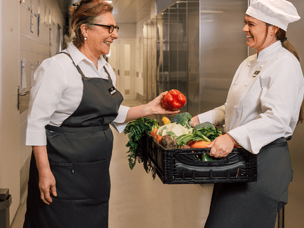 Zwei Köchinnen mit Gemüse in einer Küche lächelnd.