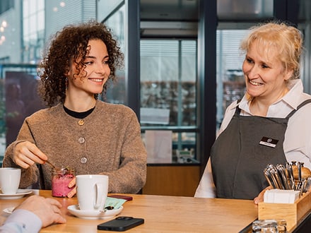 Frau lächelt und spricht mit Kellnerin in einem Café.