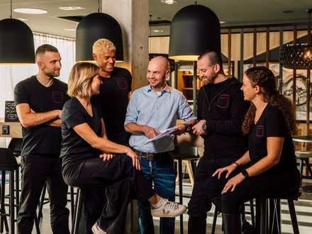 Sechs Personen, lachend, in einem modernen Café stehend.
