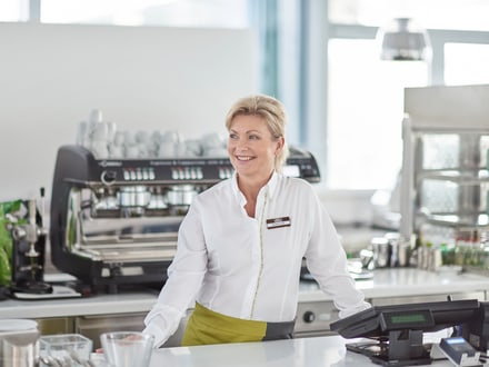 Mitarbeiterin einer Kaffeebar steht in Uniform an der Theke und lächelt.