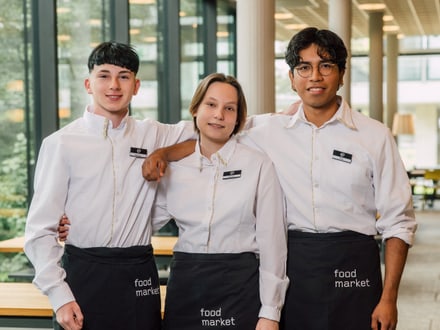 Drei Personen in weissen Hemden und schwarzen Schürzen posieren für ein Foto.