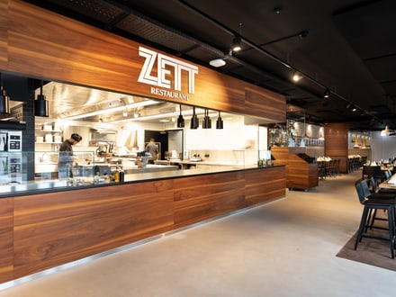 Modernes Restaurantinterieur mit Holztheke und beleuchtetem Schild 'ZEIT'.