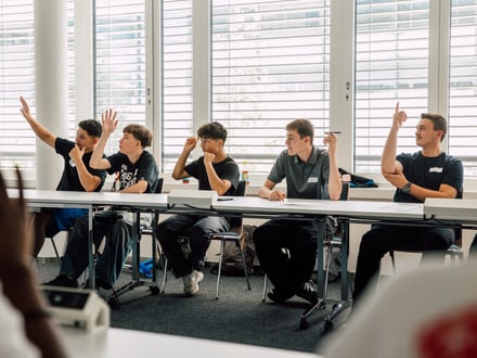 Sechs Personen heben in einem Klassenraum die Hand.