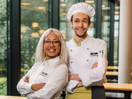 Zwei lächelnde Personen in Gastronomiekleidung stehen neben Holztischen.