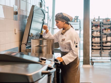 Köchin probiert Essen aus einem grossen Topf in der Grossküche.