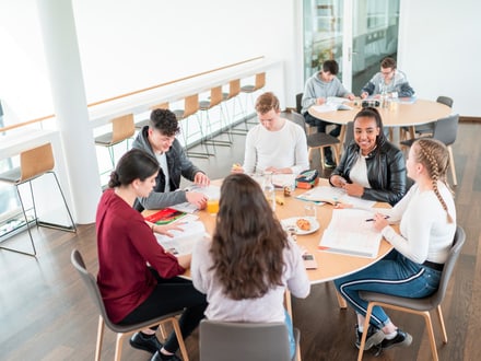 Schülerinnen und Schüler sitzen gemeinsam an einem runden Tisch in der Mensa ihrer Schule beim Lernen und geniessen dabei Snacks.