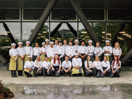 Gruppe von Köchen und Mitarbeitern vor einem Gebäude mit Glasfassade.
