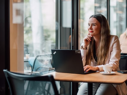 Frau im Café sitzt mit Laptop am Tisch, lächelnd aus dem Fenster schauend.