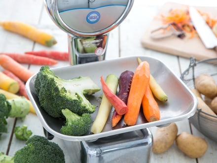 Küchenwaage mit Gemüse wie Brokkoli, Karotten und Kartoffeln auf einem Tisch.