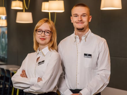 Zwei Personen in weissen Hemden und schwarzen Schürzen stehen in einem Restaurant.