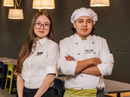 Zwei Personen in Restaurantkleidung stehen nebeneinander, eine in einem weissen Hemd und die andere in einer Kochuniform.