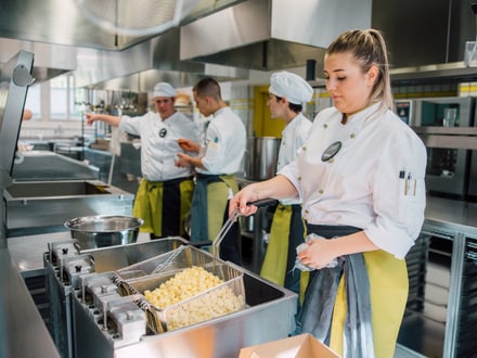 Köchin frittierende Pommes in Restaurantküche mit Kollegen im Hintergrund.",