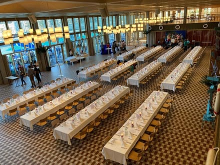 Grosser Saal mit langen gedeckten Tischen und Personen im Hintergrund.