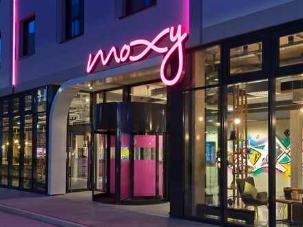 Eingang des Moxy Hotel Stuttgarts von aussen am Abend mit pink beleuchtetem Schriftzug.