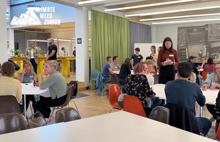 Menschen sitzen an Tischen bei Veranstaltung 'Climate Week Zurich'.