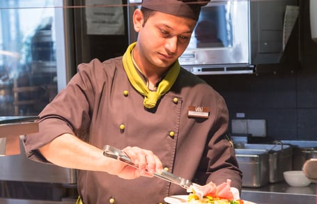 Koch mit schwarzer Blouse und Mütze mit Zange in der Hand richtet etwas auf einem Teller an. Ein Stück Fleisch ist im Vordergrund