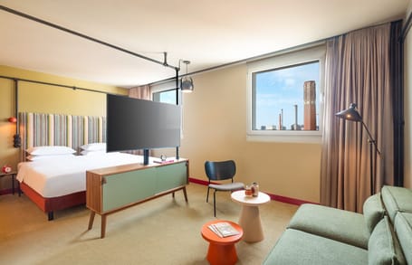 Chambre d'hôtel avec lit double, canapé, TV et vue sur le bâtiment de la ville par la fenêtre.
