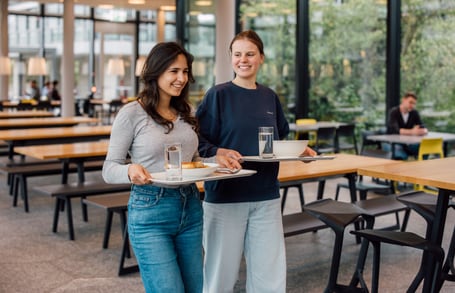 Zwei Frauen mit Tabletts in einem Speisesaal.