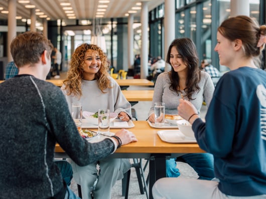 Vier Personen sitzen lächelnd in der Kantine am Tisch."  