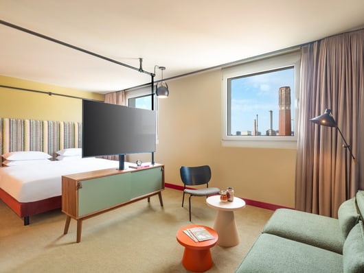 Hotelzimmer mit Doppelbett, Sofa, TV und Fensterblick auf Stadtgebäude.