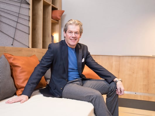 Beat Kuhn Portrait sitzt auf einer Couch in einem Hotelzimmer und trägt einen Blauen Anzug mit grauen Hosen in einer lässigen Pose