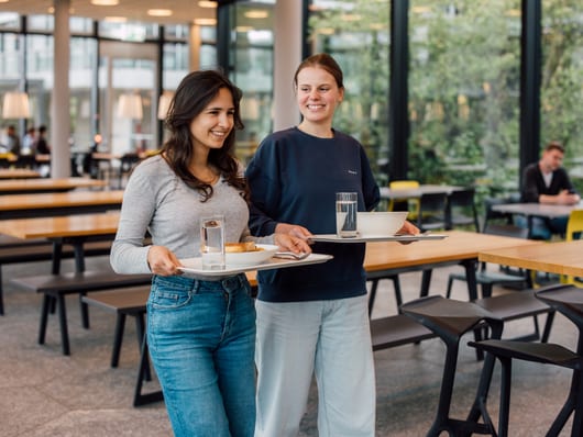 Zwei Frauen mit Tabletts in einem Speisesaal.