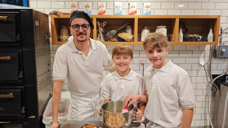 Bäcker und zwei Kinder in einer Bäckerei mit Zimtschnecken auf dem Tisch.
