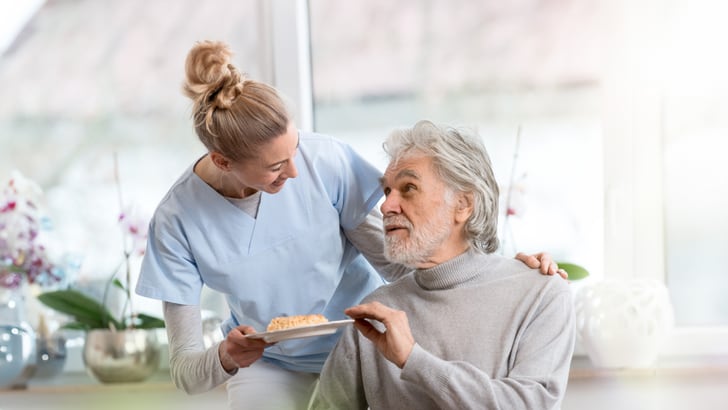 Pflegekraft in blauer Uniform serviert älterem Mann mit grauem Bart Essen.
