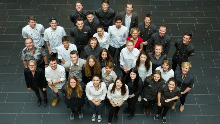 Gruppenfoto von jungen Erwachsenen in verschiedenen Berufskleidungen in einer Innenhalle, von oben aufgenommen.