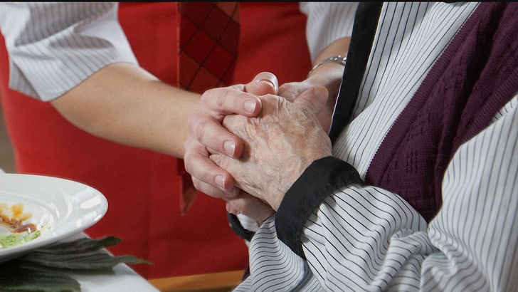Jüngere Person in roter Schürze hält Hände einer älteren Person am Esstisch.",