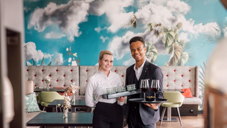 Zwei Kellner mit Tablett und Weingläsern in einem modernen Restaurant mit Wandmalerei.",