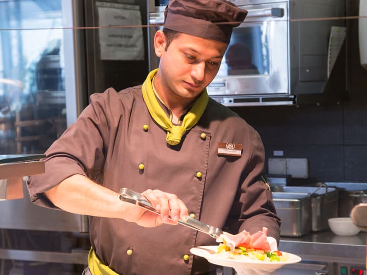 Koch mit schwarzer Blouse und Mütze mit Zange in der Hand richtet etwas auf einem Teller an. Ein Stück Fleisch ist im Vordergrund