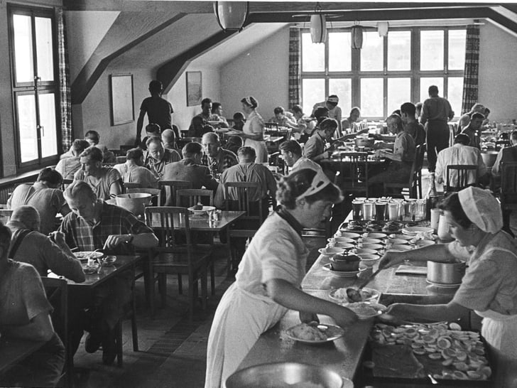 Historisches Foto eines Personalrestaurants in schwarz-weiss, wo viele Menschen beim Mittagessen sitzen und im Vordergrund an der Theke geschöpft wird.