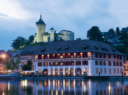 Burg und Fachwerkhaus am Abend, reflektiert im Wasser.