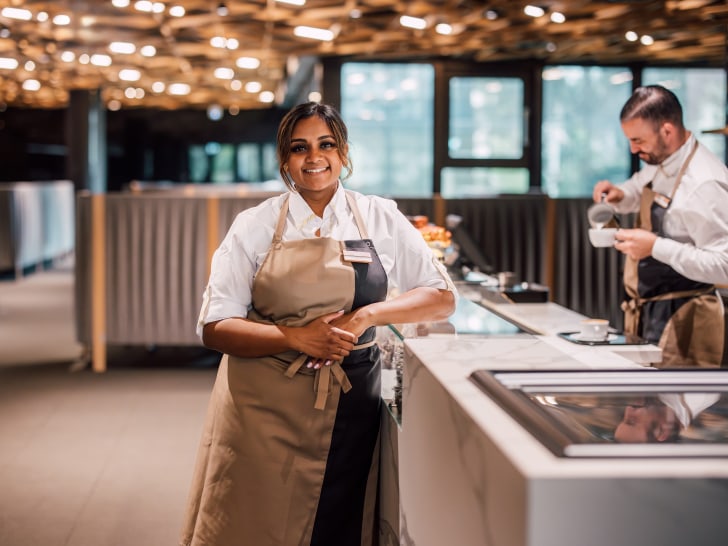 Lächelnde Kellnerin mit Schürze vor einem Restaurant-Servicecounter, Kollege im Hintergrund.