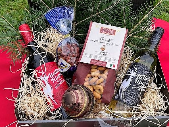 Geschenkkorb mit Weinflaschen, Nüssen, Wurst und Keksen auf rotem Tuch.