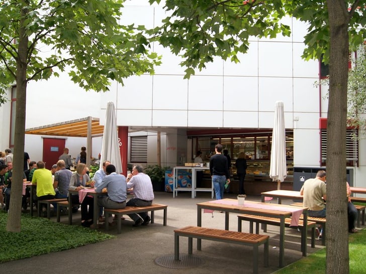 Menschen sitzen an Tischen im Freien neben einem Gebäude mit Bäumen.