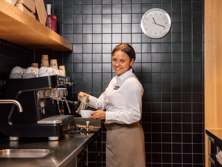 Barista macht Kaffee mit Espressomaschine, lächelnd, Wanduhr im Hintergrund.