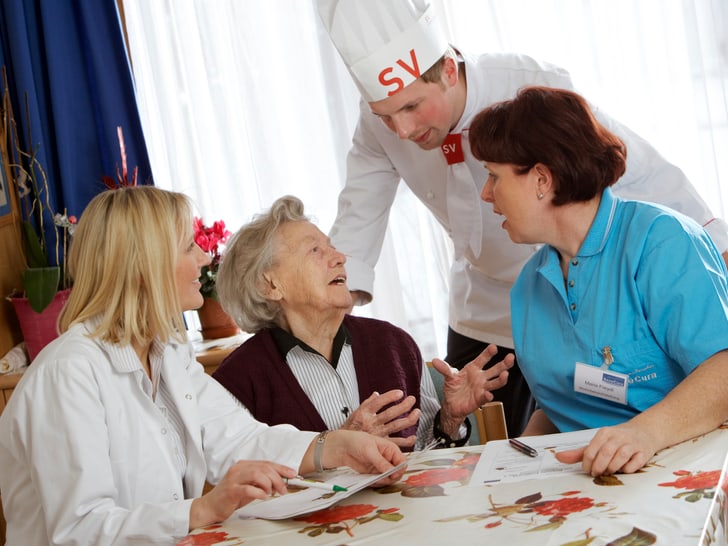Ältere Frau mit Pflegepersonal und Koch in einem Gespräch am Tisch.
