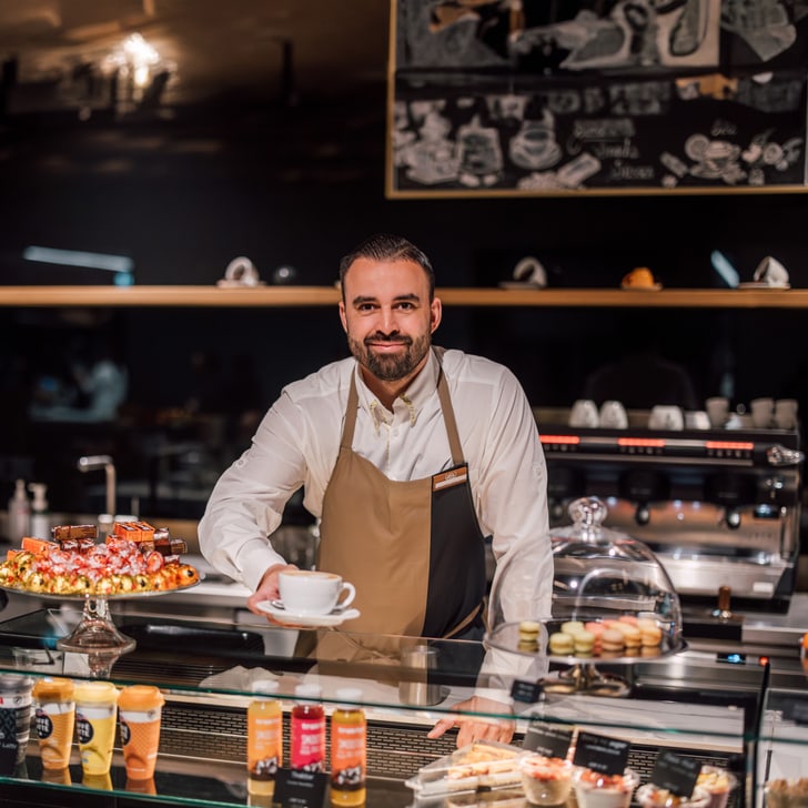 Barista mit Tasse Kaffee hinter einer Cafétheke mit Gebäck.