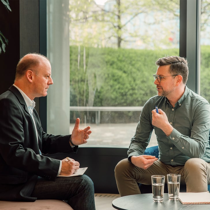 Zwei Männer im Gespräch in einem Büro mit grossem Fenster im Hintergrund.