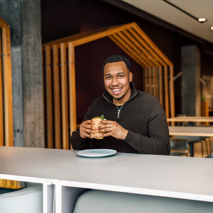 Mann sitzt an Tisch mit Sandwich und lächelt in moderner Kantine.
