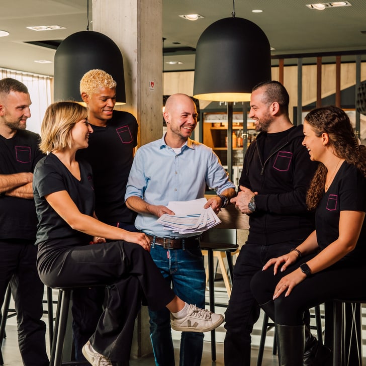 Sechs Personen lächelnd in einem Café stehend und sitzend.