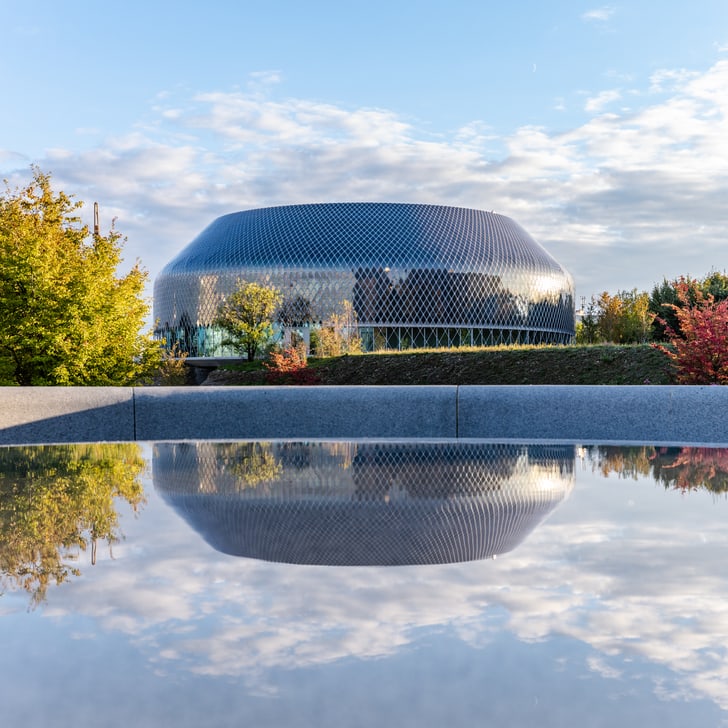 Modernes Gebäude mit Wasserreflexion und Bäumen im Vordergrund.