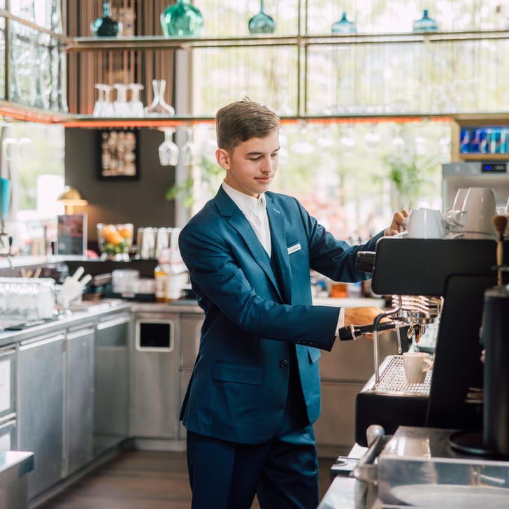 Lernender in blauem Anzug macht Kaffee an der Kaffeemaschine.